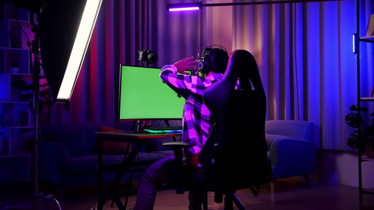 Back View Of Asian Boy Streamer Stretching After Using Green Screen Personal Computer. Live Stream On Desk Illuminated By Rgb Led Strip Light