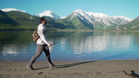 Amplia-Toma-De-Seguimiento-De-Un-Pescador-Con-Mosca-Caminando-Frente-A-Hermosas-Montañas-Cubiertas-De-Nieve-Y-Un-Lago-De-Montaña-Profundo-Y-Reflectante