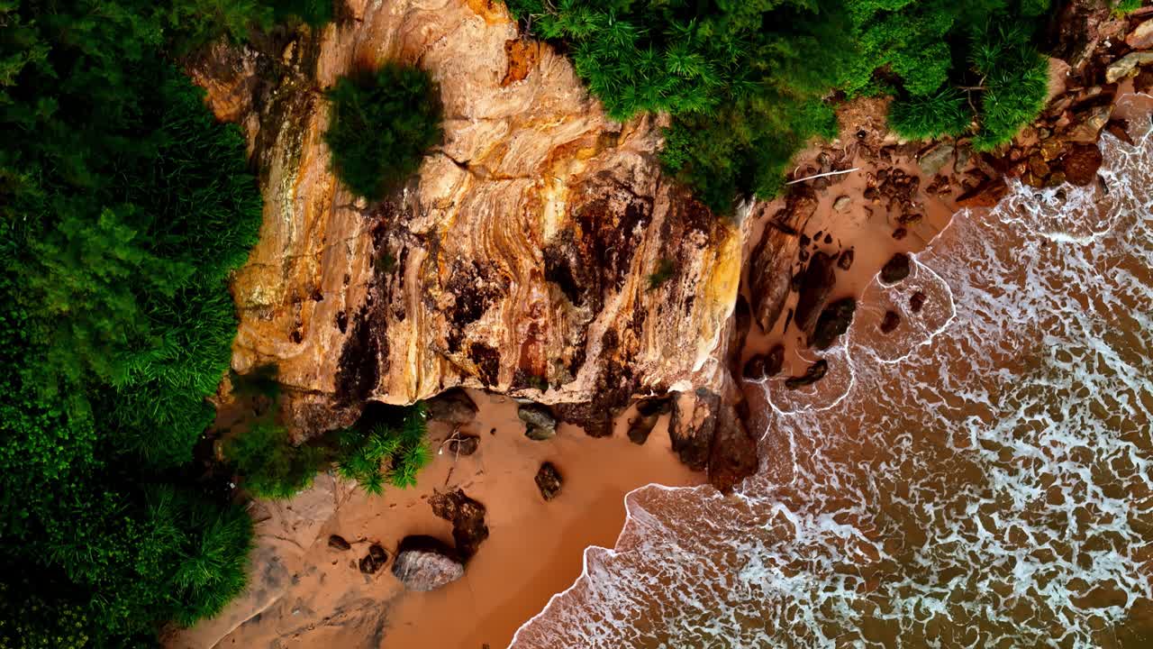 Rekaman drone udara menampilkan garis pantai batu pasir keemasan di Pantai Madiha tempat ombak pirus menghantam tebing dan tanaman hijau subur