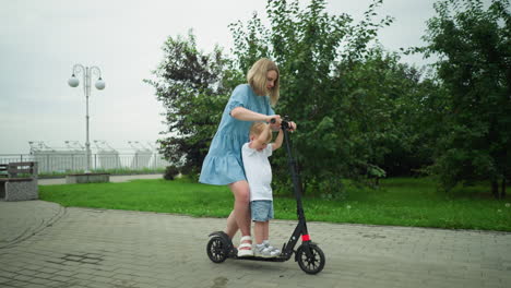 a mother and her young son ride a scooter together along a paved walkway, the mother uses her foot to push the scooter while her son holds the handlebar