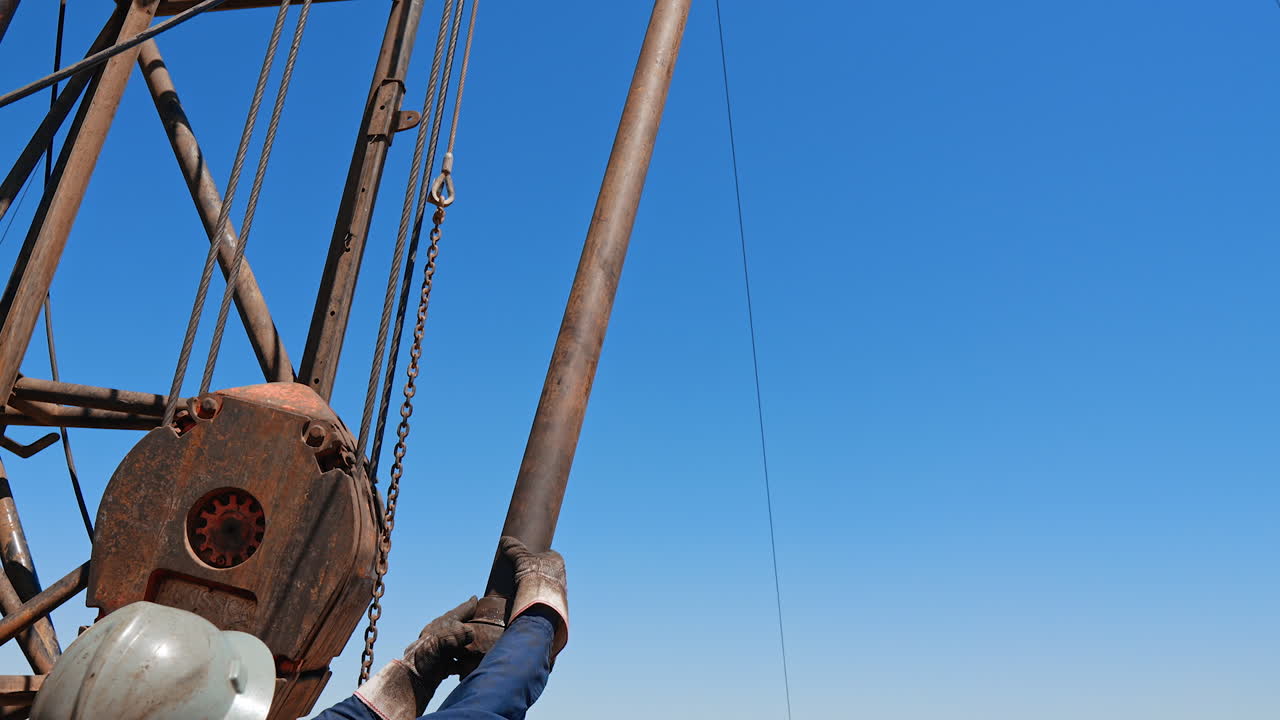 Rear view of a man wearing helmet and gloves and holding a metal pipe. Low angle view at the equipment at the site for oil production.