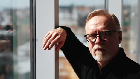 aged caucasian bearded man puts on glasses. man looks at the window on sunny day. close up.