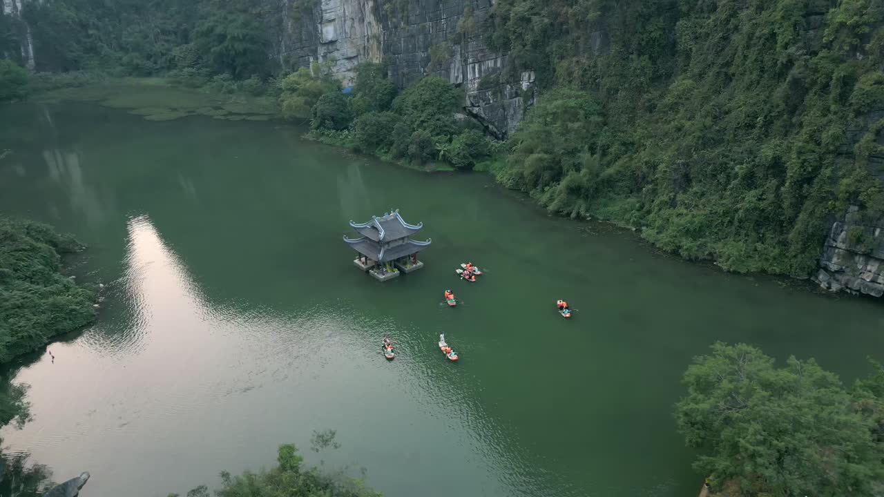 Aerial view of floating pagoda on green river, boats circling temple, lush karst mountains and cliffs form stunning natural landscape in Ninh Binh, Vietnam.