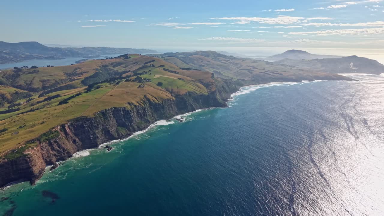High drone shot approaching green volcanic cliffs of Māori Head with waves breaking below and sunlight reflecting on the sea near Dunedin, New Zealand