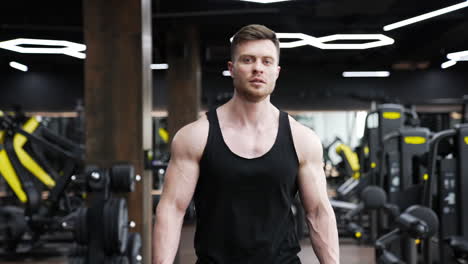 handsome athlete walks in fitness center. portrait of a young sportsman in black singlet on modern gym background. muscular bodybuilder indoors.