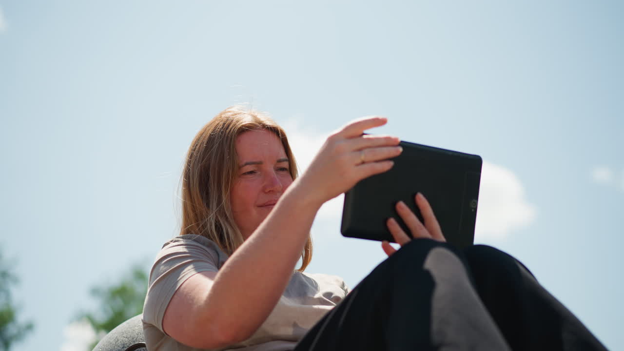 Woman sitting outdoors turning phone around with empathy while looking at screen, sunlight reflecting on her hair, serene summer day atmosphere filled with emotion