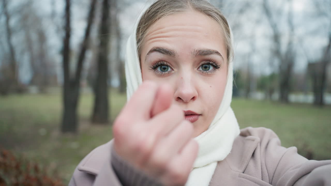 Young woman in park making gesture, white scarf and coat closeup with soft bokeh, melancholic gaze shifts to playful beckon, cold overcast atmosphere, intimate invitation toward camera