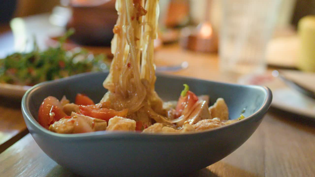 Hands mix spicy noodle salad with tomatoes and pork skin in warmly lit restaurant setting