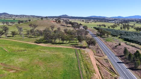 aerial drone footage tracks above a rural road winding through green fields, scattered trees, and rolling hills under bright midday sunlight
