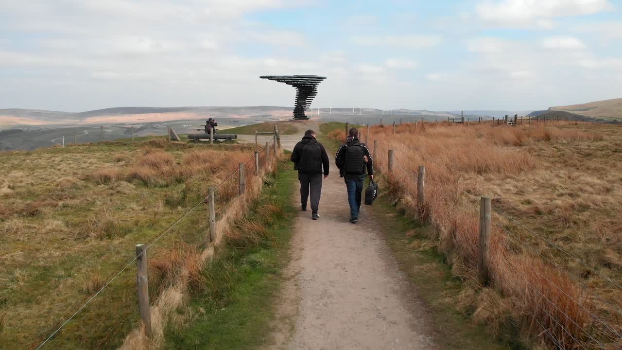 Wide shot of two hikers walking on a rural path toward the Singing Ringing Tree sculpture