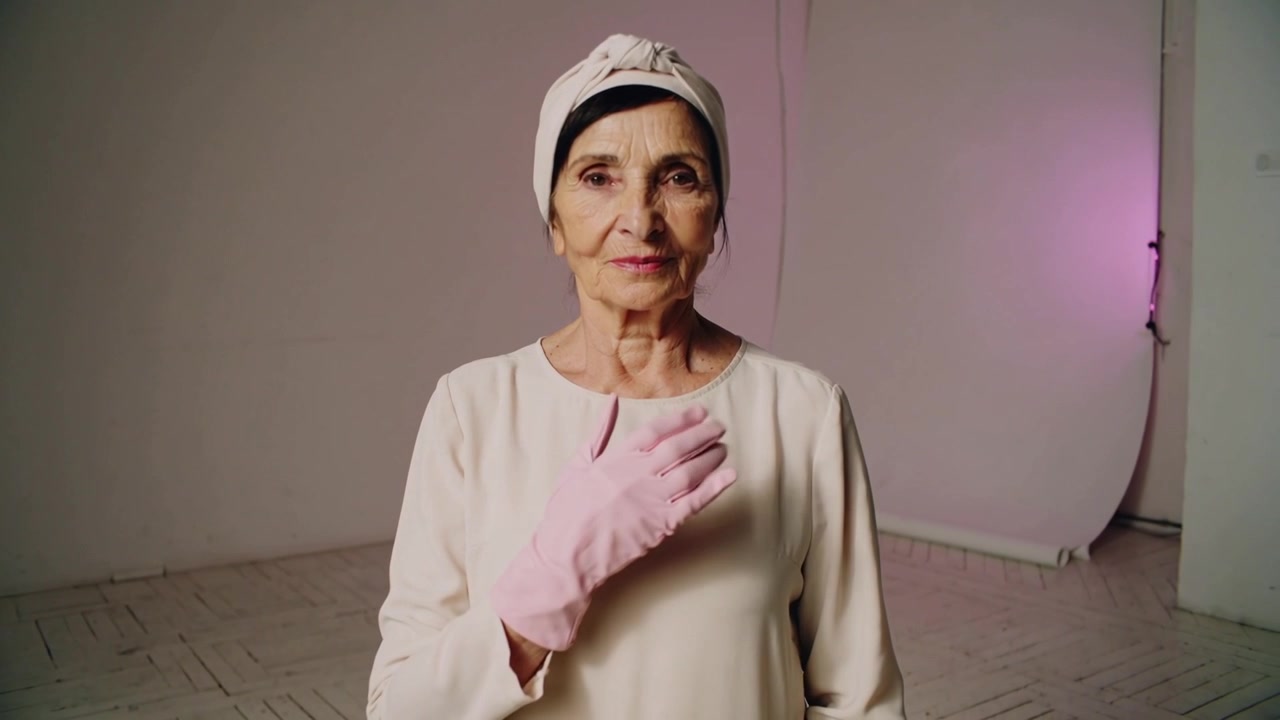Donna anziana in posa che si tocca il petto dopo una sequenza iniziata in studio con guanti rosa e sciarpa