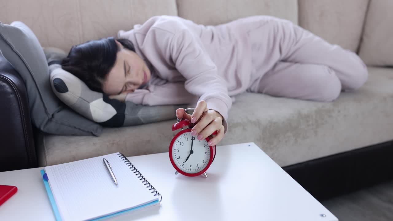 若い女性がソファで眠り、鳴っている目覚まし時計を止める