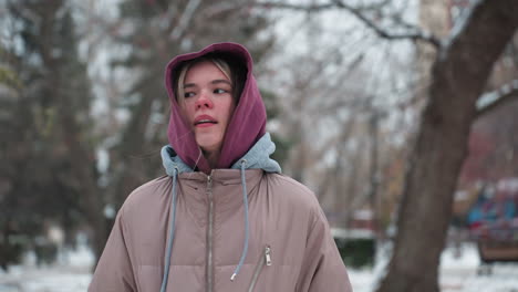 girl feeling cold while jumping with thoughtful facial expression in winter park, dressed in hoodie and coat, she appears lost in thought against a blurred snowy background of trees and benches