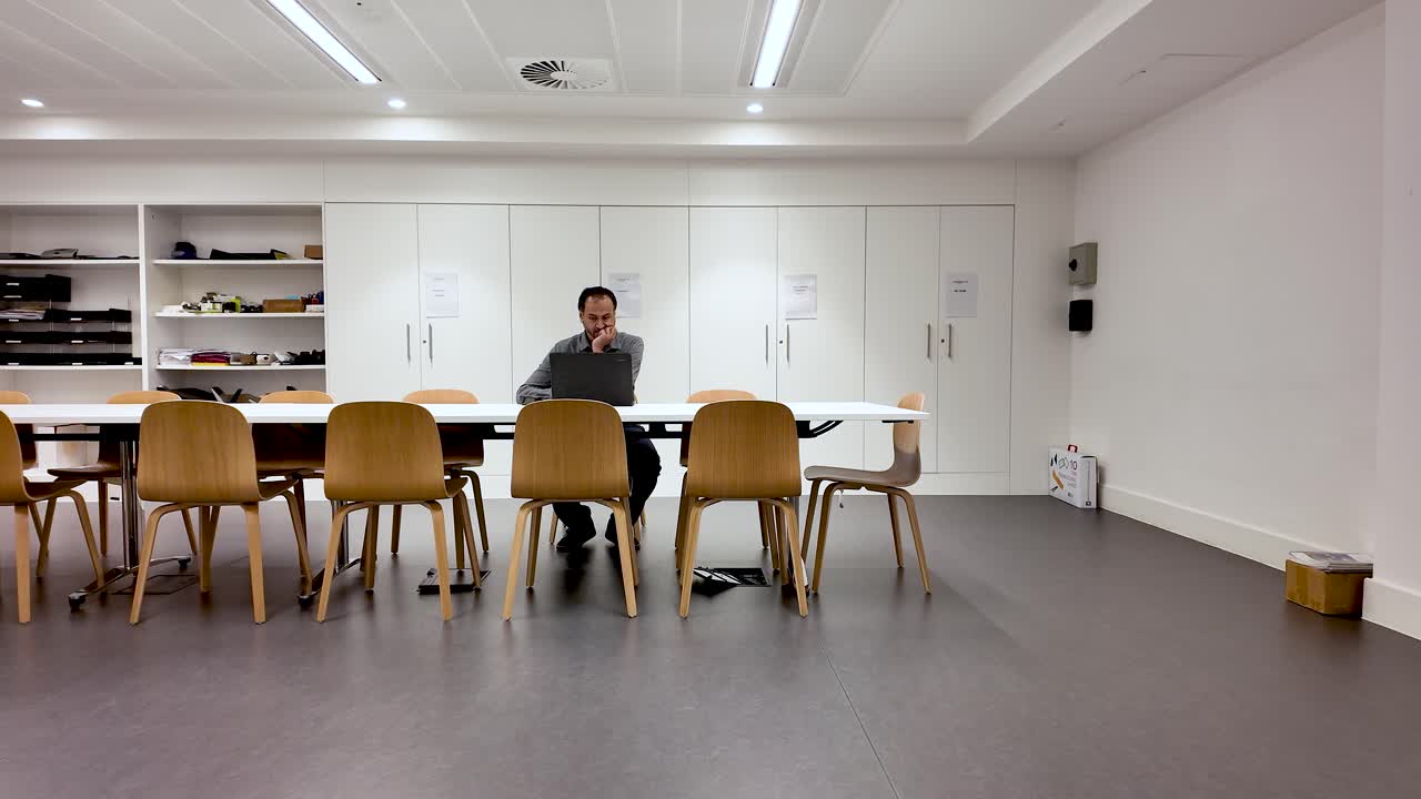 South Asian man working on laptop at a long conference table in an empty office