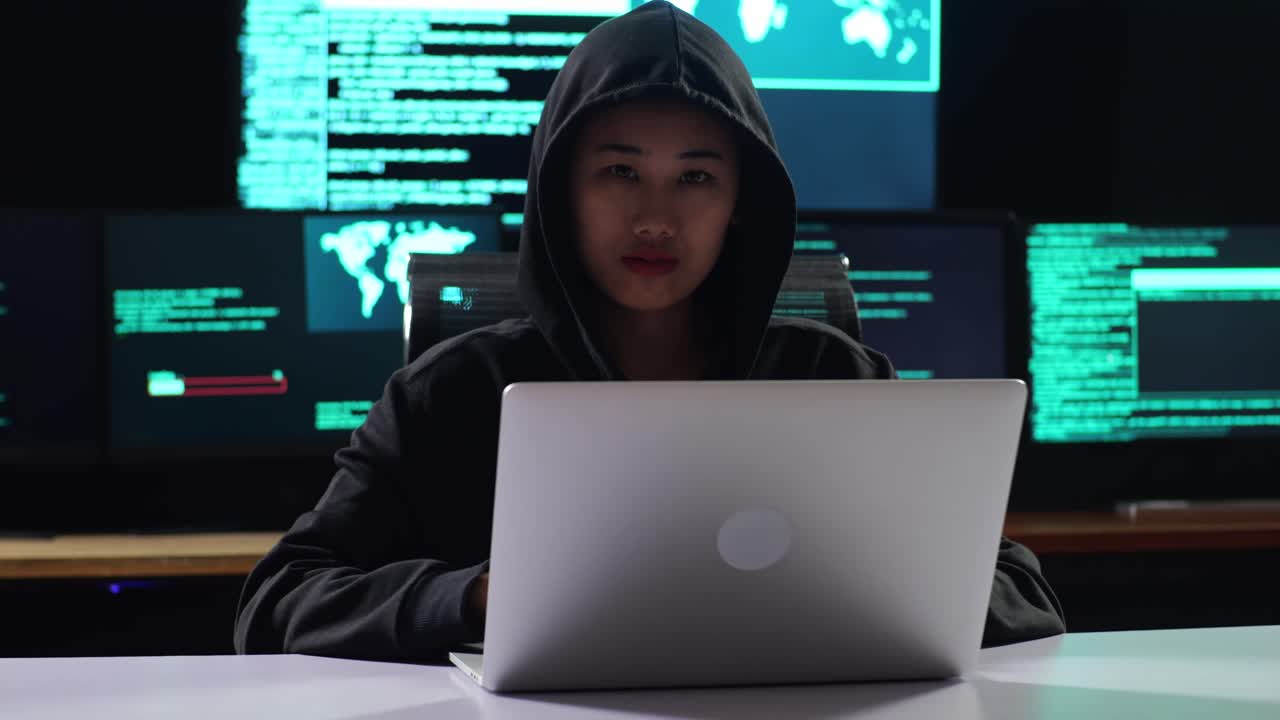 어두운 사무실 방에서 여러 모니터의 디스플레이 스크린에 지도와 데이터를 가진 노트북에서 작업하는 동안 카메라에 손바닥을 보여주는 후드를 입은 아시아 여성 해커