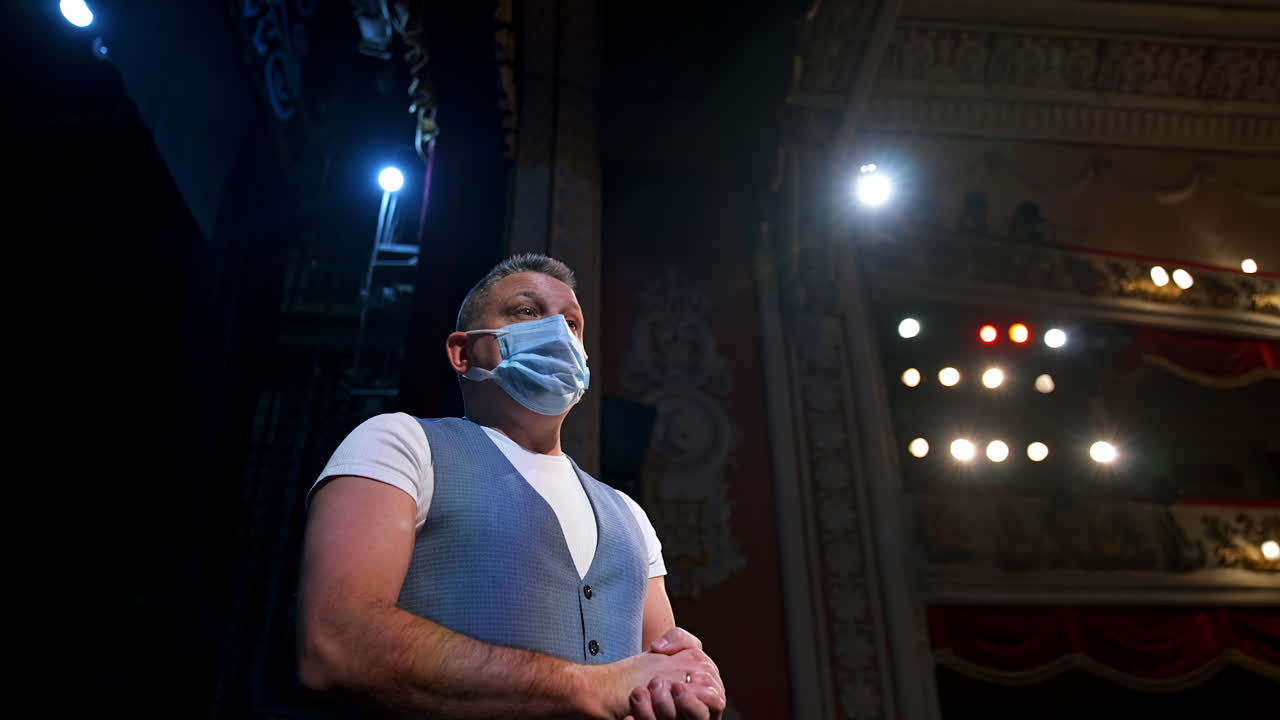Actor in medical mask talking. Man is standing on stage in theater at spotlights and speaking during pandemic. View from below.