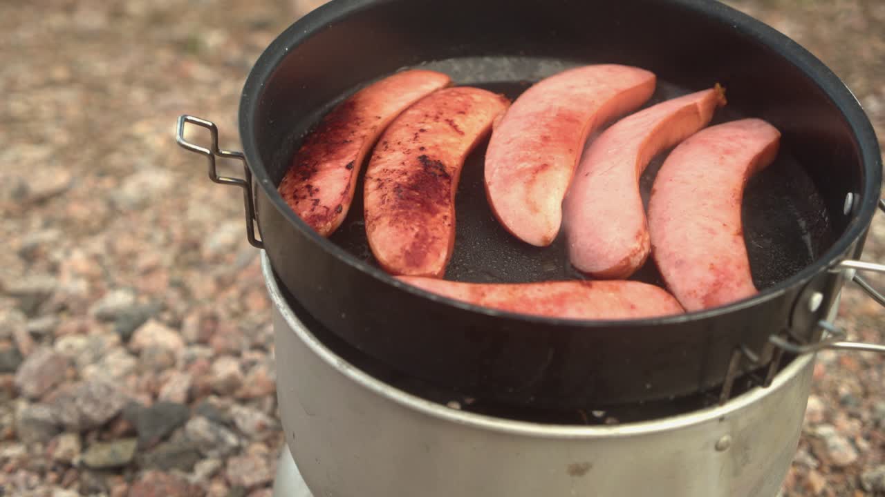 Tilt up from camp stove on rocky ground to sausages frying in hot pan