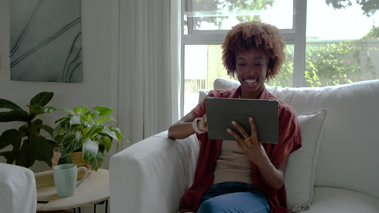 African American woman watching tablet content, leaning forward, tapping, laughing on couch