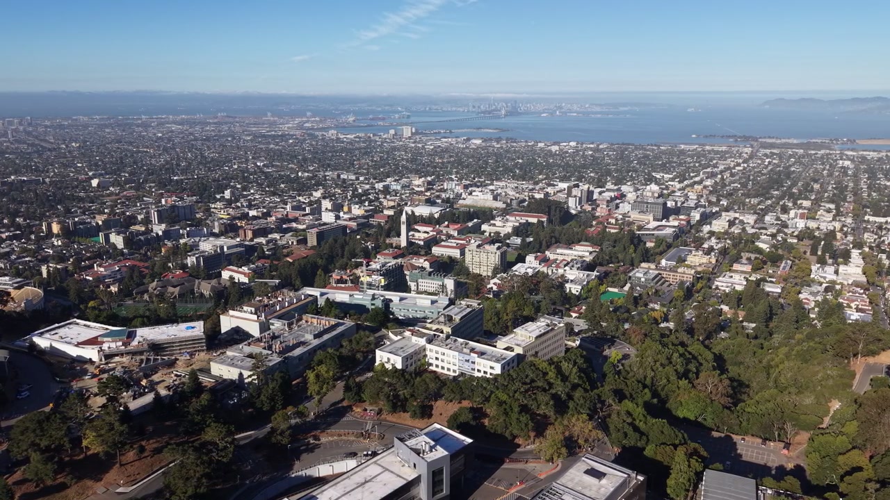 Ampia vista aerea che rivela lentamente il campus dell'Università della California, Berkeley, con lo skyline della città, la Baia di San Francisco e l'iconico Bay Bridge visibili sullo sfondo soleggiato e velato