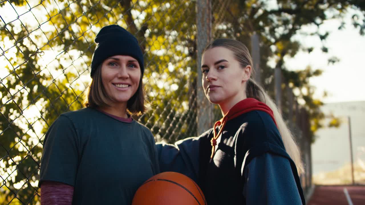 retrato de dos chicas felices en ropa deportiva con una pelota naranja de baloncesto en un campo deportivo callejero por la mañana en un verano soleado