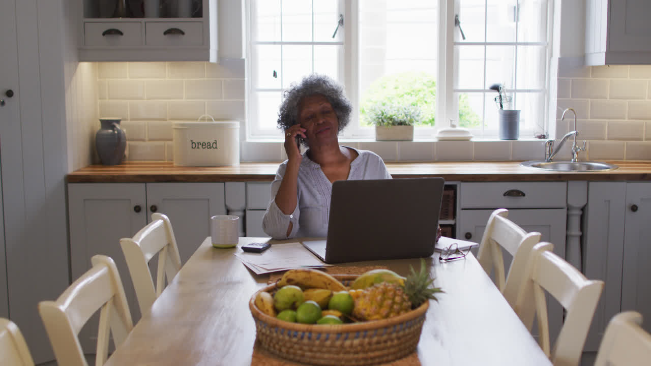 mujer afroamericana mayor usando una computadora portátil y hablando en un teléfono inteligente en casa