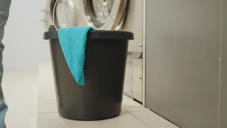 Down view black bucket with blue towel set beside stainless washing machines in bright laundromat, woman bends to lift container from tiled floor, starting cleaning routine, focus on utility gear
