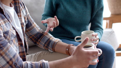 happy biracial couple sitting on sofa, drinking coffee and talking at home, in slow motion