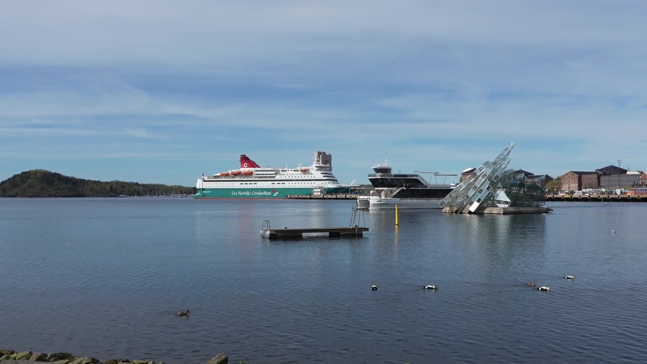 Håndholdt optagelse af Oslo Fjords havn med både og ænder under klar blå himmel, med skulpturen She Lies, fortøjet krydstogtskib, stille vand og fjerne bygninger