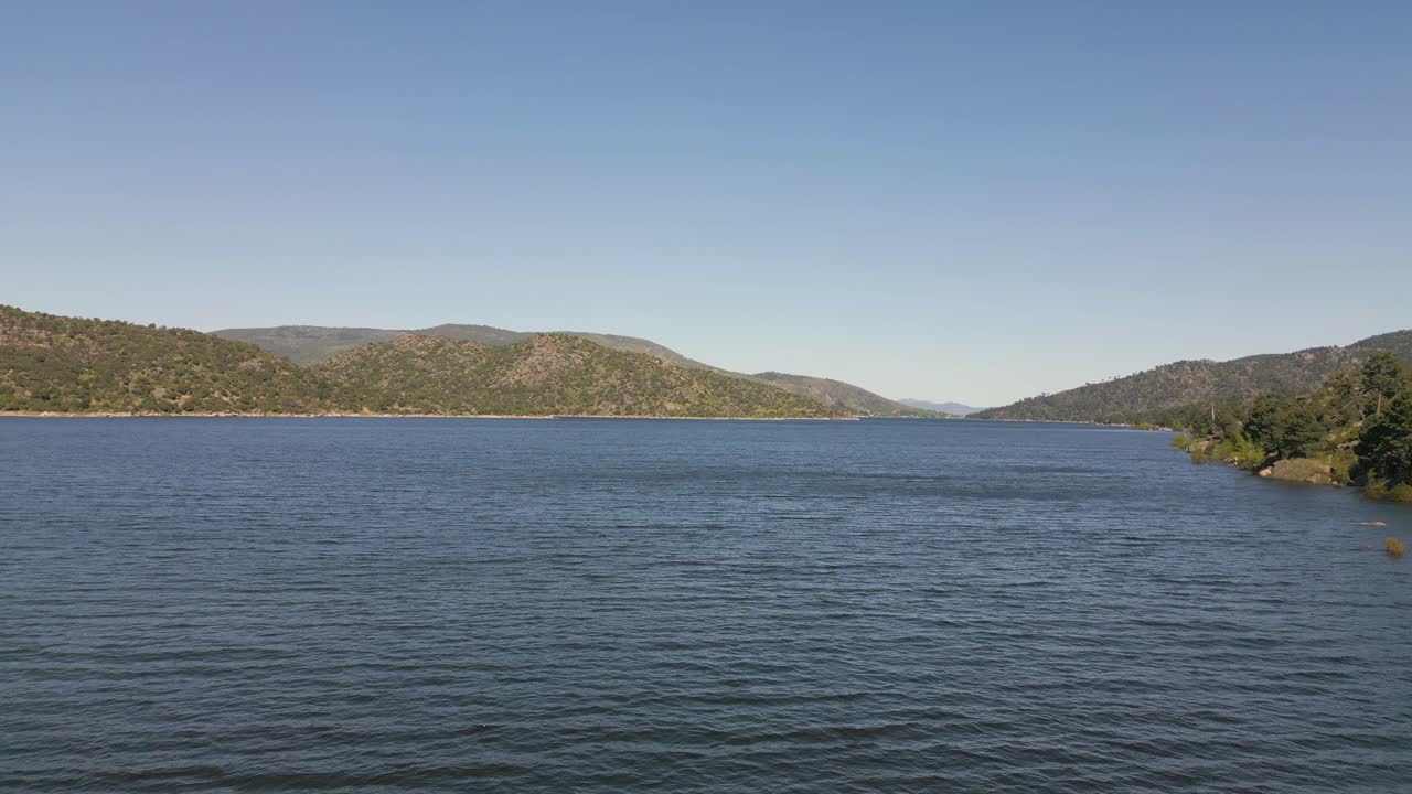 Descending drone flight over a large reservoir with calm water, surrounded by a beautiful mountainous landscape under a clear blue sky on a lovely day. Peaceful and natural atmosphere.