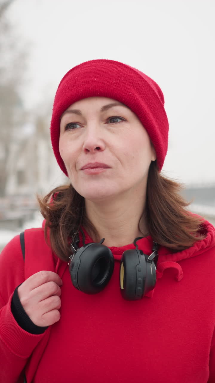 Close-up of woman walking on interlocked pathway holding bag over shoulder with calm expression near iron railing, background shows blurred lamp posts and snowy trees in foggy winter atmosphere