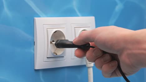 a woman inserts a black narrow electric plug into a white electrical outlet mounted on a blue wall, bathroom, interfacing, docking, wiring