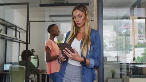 caucasian businesswoman leaning in doorway using tablet, smiling to camera in busy office