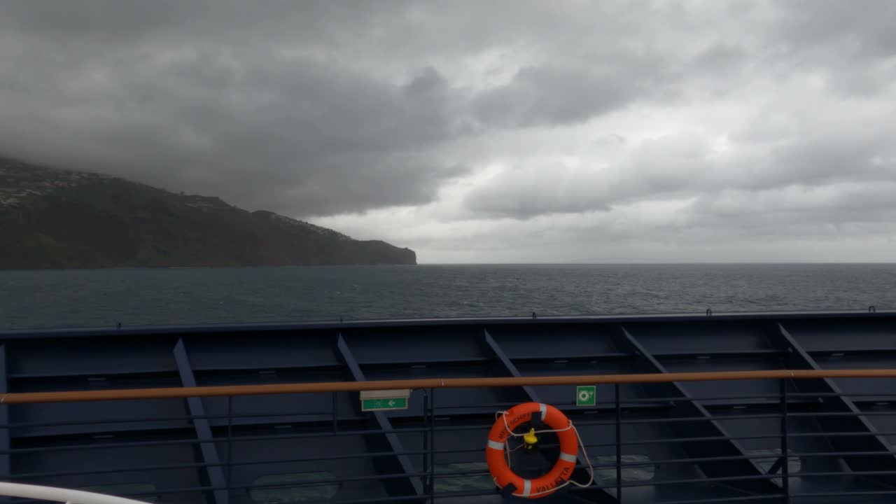 Dramatischer Zeitraffer vom Fährdeck, zeigt stürmischen Atlantik, dunkle Wolken über den Klippen Madeiras und einen Rettungsring. Stimmungsvolle Meereslandschaft nahe Funchal, Portugal