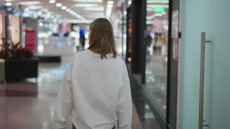 back view of a shopper in a white top walking through a shopping mall, looking around amid colorful lights and a blurred background with unclear objects