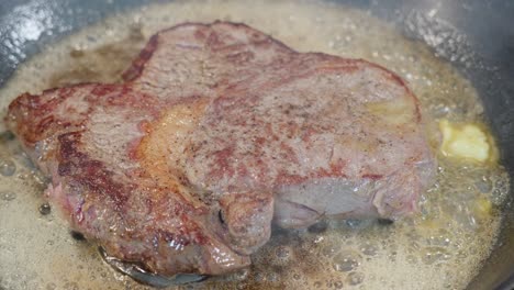 Closeup-of-entrecote-steak-frying-in-a-hot-pan,-added-butter-melting-and-bubbling-with-smoke-rising