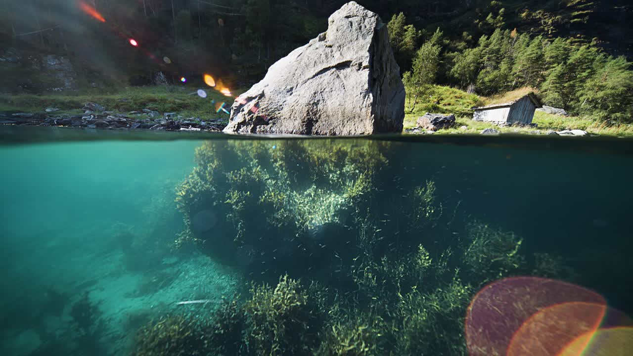 The calm waters of Geiranger Fjord reveal the submerged half of a rock, with greenery and rocky terrain onshore