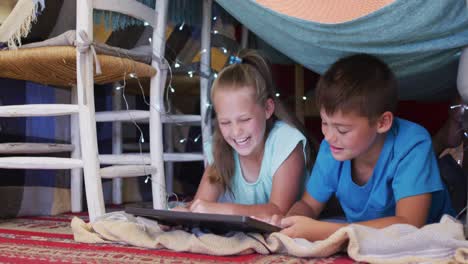 caucasian brother and sister smiling while using digital tablet under the blanket fort at home