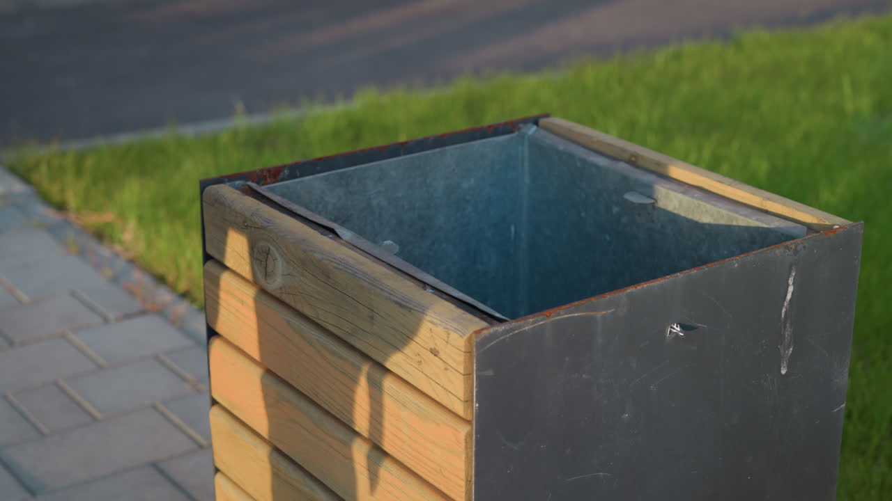 close up of outdoor metal and wooden waste bin with subtle human shadow visible on surface as unidentified person throws object into bin under warm sunlight near grassy urban pavement
