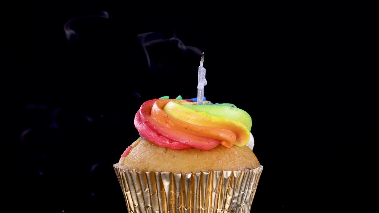 Un cupcake con glassa arcobaleno e una candela che viene spenta, rilasciando una scia di fumo visibile su uno sfondo nero