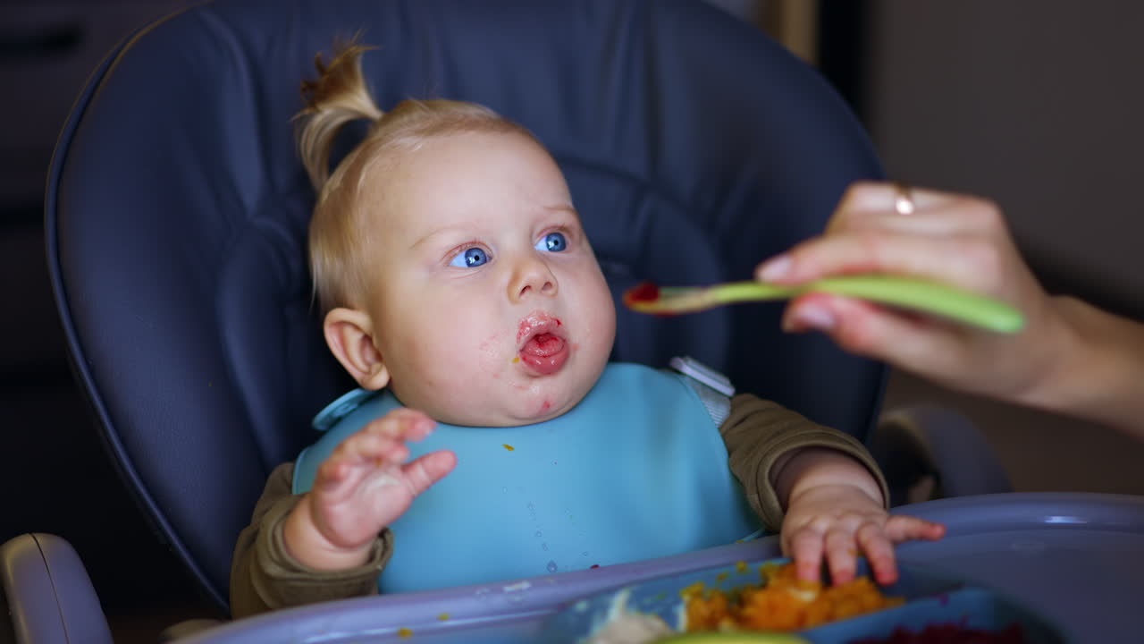 Lovely Caucasian baby boy sitting in high chair is fed from spoon. Mother giving pureed veggies to an infant. Close up.