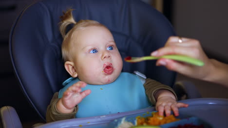 lovely caucasian baby boy sitting in high chair is fed from spoon. mother giving pureed veggies to an infant. close up.