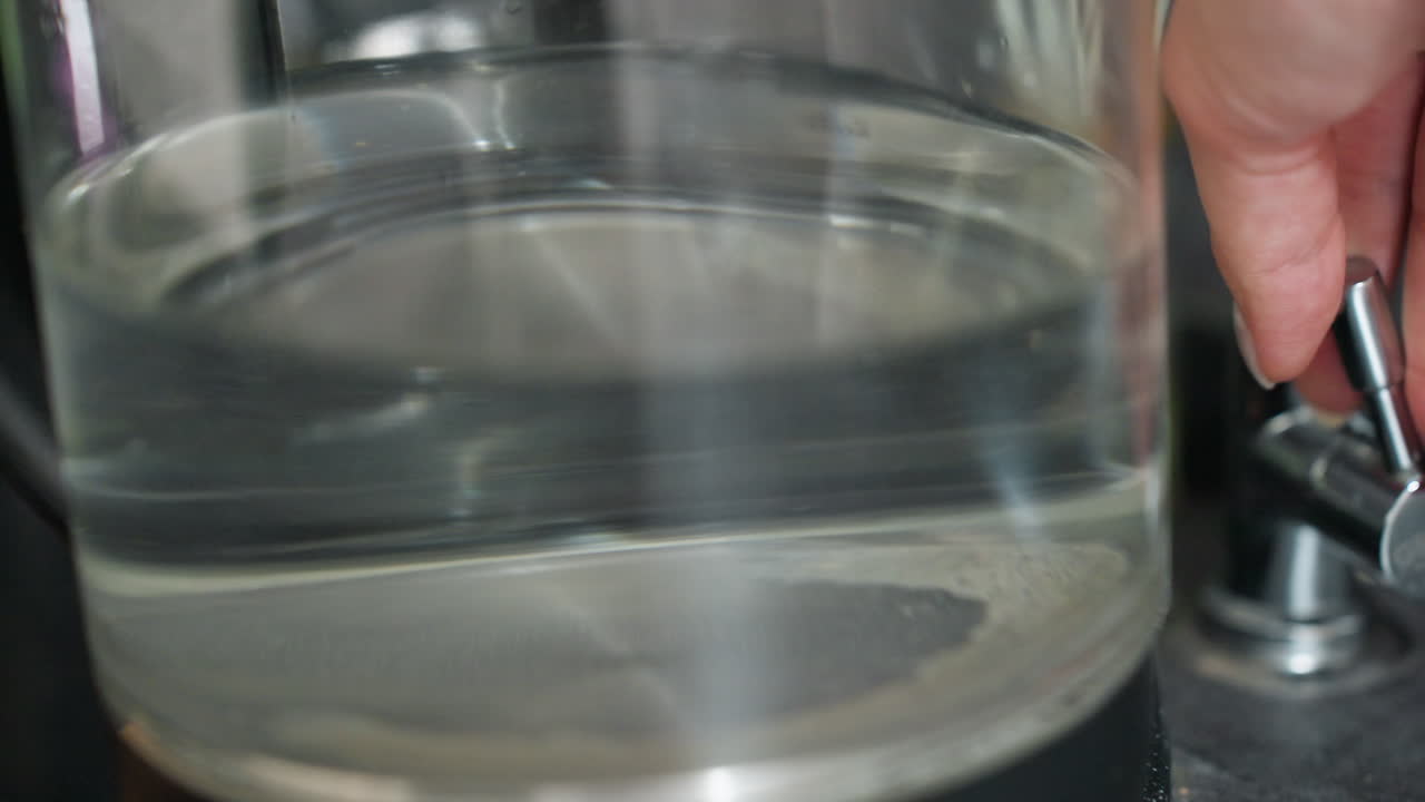 Aggiunta accurata di acqua a una brocca di vetro, mani ferme versano acqua in un contenitore trasparente ripreso visivamente, trasferimento cauto di acqua dalla brocca al recipiente in una scena di cucina organizzata