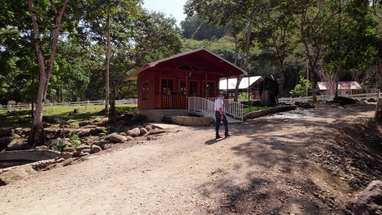A man walks towards a red wooden house in a lush, natural environment