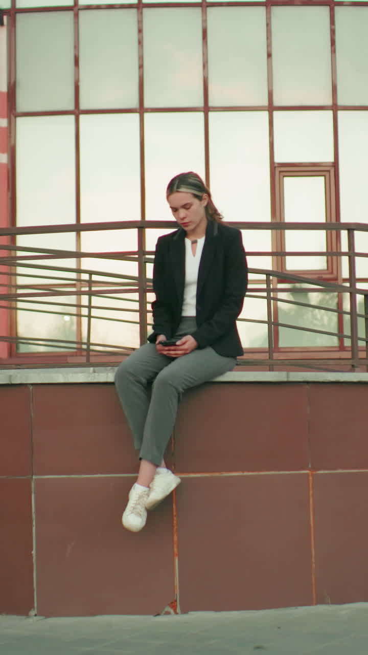 Woman seated alone on fence in solitude with legs crossed at ankle, looking at phone in hand, appearing thoughtful in front of modern building with lamp posts and railings on tiled platform