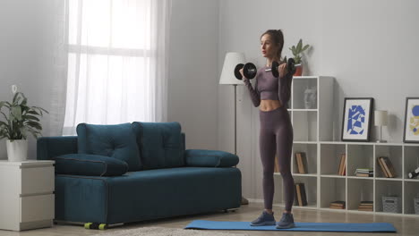 young sporty woman is training with dumbbells standing in living room at daytime full-length portrait of sportswoman in modern interior