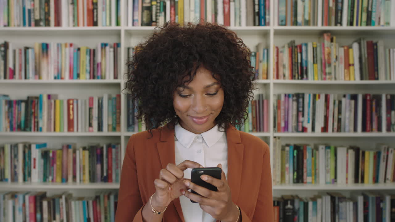portrait of stylish young african american business woman intern texting browsing online using smartphone technology in library bookshelf background