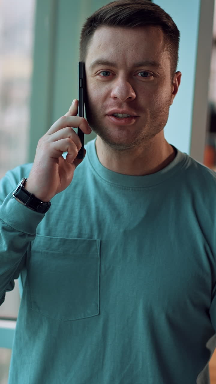 Fit mid-aged man looking straight into camera speaking on the phone. Businessman having phone conversation standing near the window. Female sitting at desk at backdrop. Vertical video