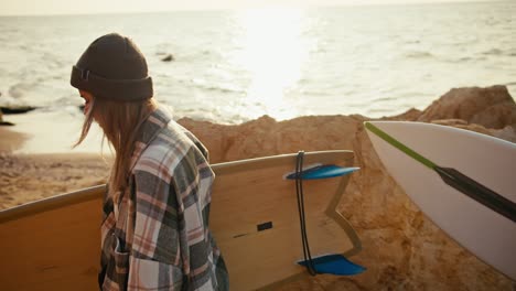 Close-up-shot-of-a-brunette-guy-in-a-white-T-shirt-and-a-blonde-girl-in-a-checkered-shirt-walking-along-the-rocky-seashore-and-carrying-surfboards-in-the-morning-at-sunrise