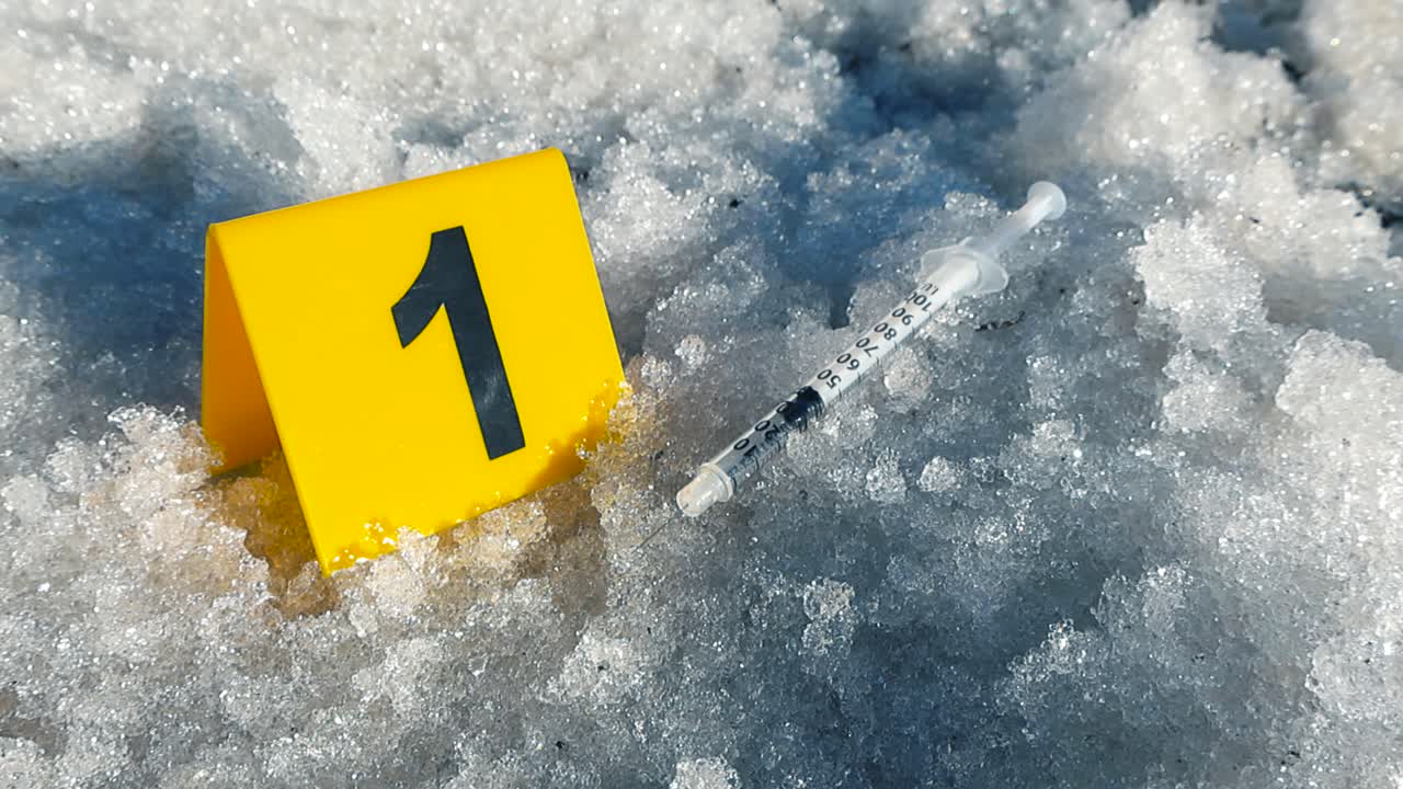 Yellow forensic marker indicating a used syringe as evidence on the snowy ground at a crime scene, symbolizing illegal drug use, danger, and police investigation in an urban environment
