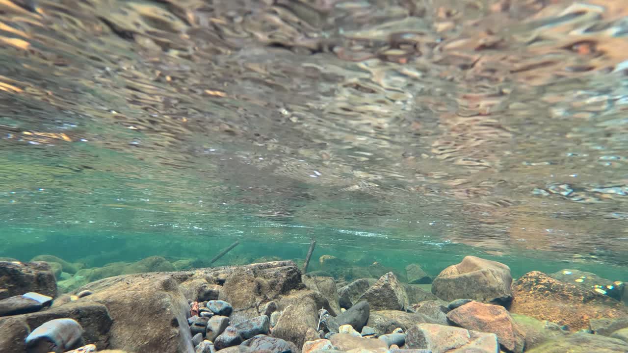 Camera submerges beneath a clear mountain stream, revealing underwater rocks and flowing water under a stone bridge on the Isle of Skye